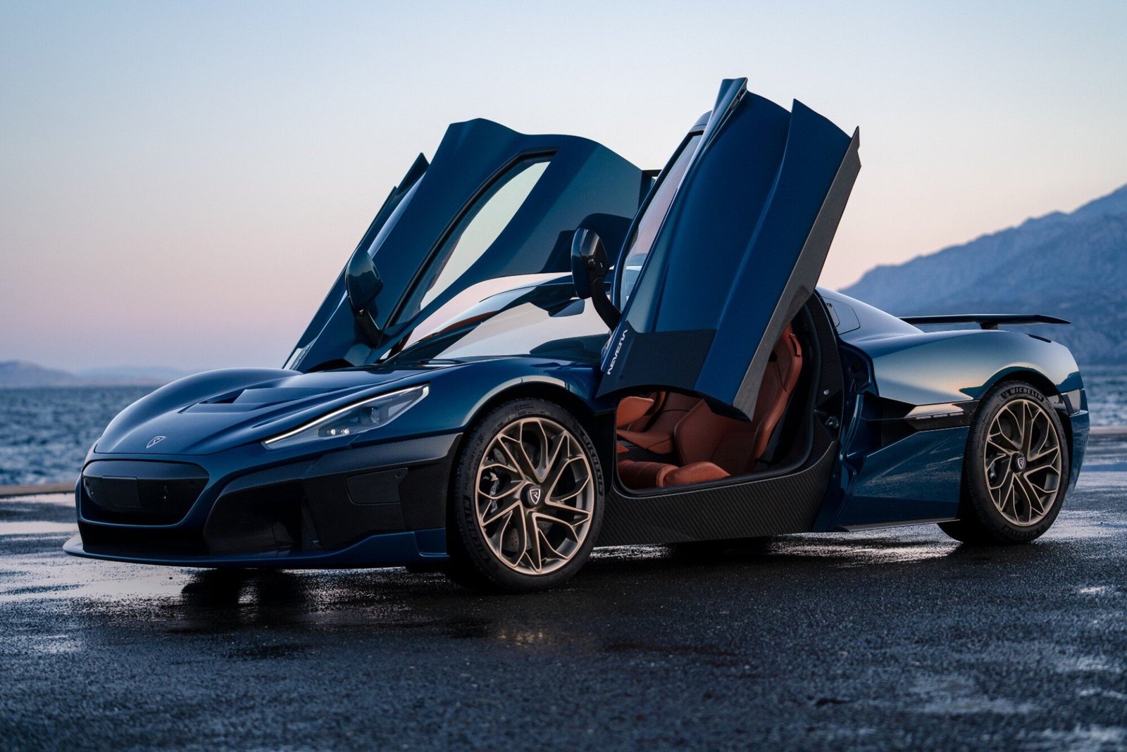 A photo of a blue Rimac Nevera parked in front of a sunset with its doors up.